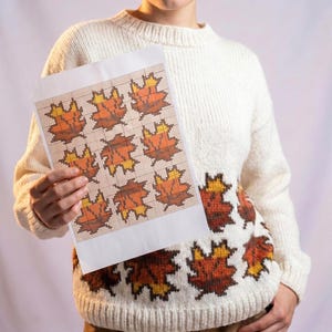 May include: A cream-colored long-sleeve knit sweater featuring a repeating pattern of autumn leaves in shades of orange, brown, and yellow. A white paper sheet is held up, displaying a grid-based cross-stitch pattern of the same leaf design.
