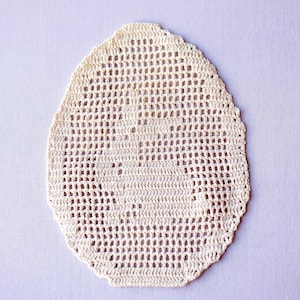 May include: An egg-shaped off-white crochet doily featuring a central motif of a sitting rabbit. The piece is crafted with a fine thread in a grid-like openwork pattern, with denser stitches forming the silhouette of the bunny in the middle.