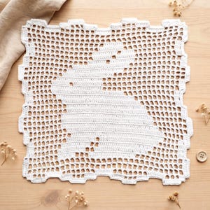 May include: A square white crochet doily featuring a central silhouette of a rabbit created with dense stitching against an open grid background. The edges of the textile are scalloped and textured, showcasing intricate needlework and yarn detail.