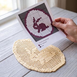 Op de afbeelding: Een beige ovaal gehaakt kleedje met een centraal konijn-silhouetpatroon, vergezeld van een geprint papieren rasterdiagram dat de bijbehorende steekinstructies toont met een legenda voor lege vierkantjes, blokken en gebieden zonder steken.