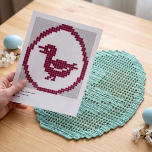 May include: A printed paper pattern featuring a pixelated dark red duck inside an egg-shaped outline, held next to an oval-shaped light mint green crocheted doily with a textured, open-work stitch pattern and scalloped edges.