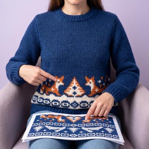 May include: A dark blue knit sweater featuring a repeating pattern of orange foxes, white trees, and geometric shapes. The sweater has a crew neckline and long sleeves. A printed paper pattern matching the sweater design is held in front.