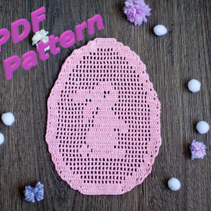May include: A pink crocheted doily in the shape of an egg on a wooden surface. The doily has a delicate pattern and is surrounded by small white pom-poms. The text "PDF Pattern" is in pink 3D letters in the top left corner of the image.