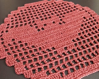 May include: A handmade, dusty rose crochet doily with a geometric openwork design. The square doily features a scalloped edge and a textured, raised central panel. The open weave allows the surface beneath to be visible, creating a delicate, vintage aesthetic.