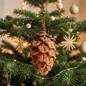Peut inclure: Une décoration en forme de pomme de pin crochetée avec des écailles en fil marron clair superposées et une partie supérieure marron foncé. L'ornement est suspendu par une fine boucle de ficelle marron, présentant un design texturé et artisanal.