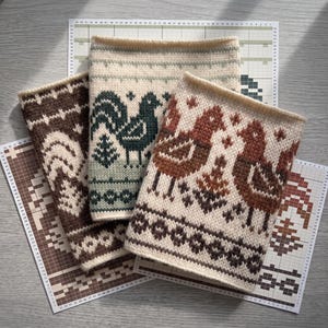May include: Three knitted neck warmers featuring fair isle patterns of birds and geometric shapes. One is brown and cream, one is sage green and cream with a dark green bird, and one is rust orange and cream. Each rests on a printed grid pattern.
