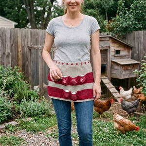 May include: A beige crochet egg gathering apron featuring two horizontal rows of deep red pockets, each designed to hold a single egg. The apron is secured around the waist with matching beige ties and is accented with a small row of red star motifs.