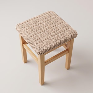 May include: A square wooden stool featuring a light beige, hand-knitted seat cover. The cover displays a textured, repeating geometric square pattern. The stool has four straight, light-toned wooden legs connected by horizontal support bars.