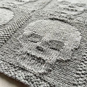 May include: A light gray knitted textile featuring a repeating pattern of raised skull motifs. The fabric displays a textured knit construction where the skull shapes are formed by smooth stockinette stitches against a background of seed stitches.