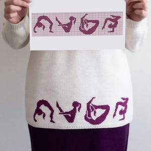 May include: A white sweater featuring a purple graphic pattern of four distinct yoga poses along the bottom hem. The same four yoga poses are displayed on a paper grid held above the sweater, showing the design layout for the knitted garment.
