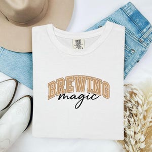 May include: A white t-shirt with the words "BREWING magic" in a brown, arched font. The word "magic" is in a cursive font. The shirt is folded and placed on a light background with a hat, jeans, and boots.