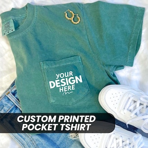 May include: A teal green t-shirt with a pocket on the left side. The pocket has the text "YOUR DESIGN HERE" printed on it. The t-shirt is laying on a white surface with a pair of jeans and white sneakers.