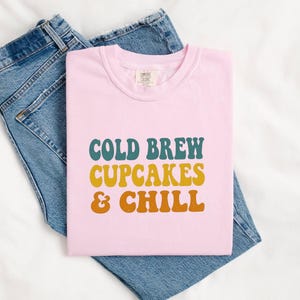 Op de afbeelding: Een lichtroze t-shirt met de tekst "COLD BREW CUPCAKES & CHILL" in turquoise, geel en oranje letters. Het shirt ligt op een blauwe spijkerbroek.
