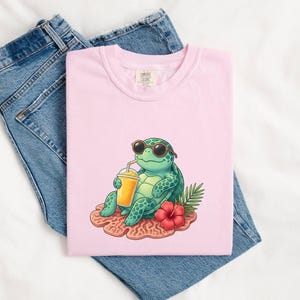 May include: A light pink t-shirt featuring a cartoon turtle wearing sunglasses and drinking from a juice cup. The turtle is sitting on a coral-like surface with a red flower and green leaves. The shirt is laid on top of blue jeans.