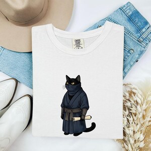 May include: A white crew neck t-shirt featuring a graphic of a black cat dressed as a ninja in a dark blue robe and mask, holding a small scroll. The shirt has a Comfort Colors label at the inner neckline and is displayed with a hat and jeans.