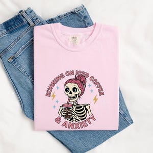 Peut inclure: T-shirt rose clair avec un motif de squelette buvant du café glacé, avec le texte "RUNNING ON ICED COFFEE & ANXIETY". Le t-shirt est plié sur un jean bleu.