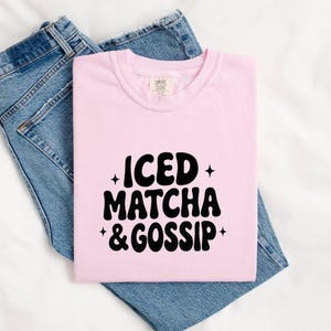 May include: A pale pink t-shirt featuring the text "ICED MATCHA & GOSSIP" in black, retro font. The t-shirt is placed on top of a pair of blue denim jeans.
