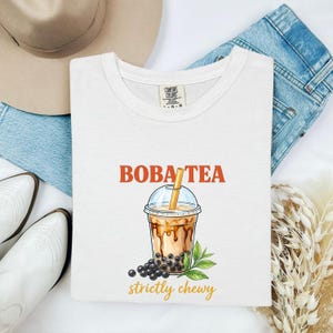 May include: White t-shirt with a crew neck featuring the words "BOBA TEA" in orange. Below the text is an illustration of a boba tea drink with a straw, boba pearls, and the words "strictly chewy" in gold.