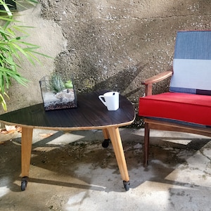 Peut inclure: Une table basse triangulaire avec un plateau en bois foncé et des pieds en bois clair. La table est équipée de roulettes. Un terrarium et une tasse blanche sont posés dessus. Un fauteuil en bois avec un coussin rouge est visible en arrière-plan.