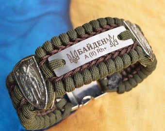 Könnte beinhalten: Ein grünes und braunes Paracord-Armband mit einer silbernen Metallplatte, auf der "БАЙДЕН A (II) Rh+" eingraviert ist, und einem silbernen Metallanhänger mit einem religiösen Bild.