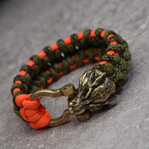 Puede incluir: Un brazalete de paracord verde y naranja con un cierre de cabeza de lobo. El cierre está hecho de latón y tiene un diseño detallado.