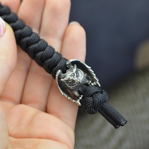 Peut inclure: Un bracelet en paracorde noir avec un charme d'hibou en argent. Le hibou est détaillé avec des ailes et une expression féroce.