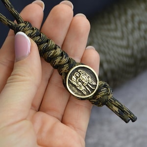 Könnte beinhalten: Ein grünes und schwarzes Paracord-Armband mit einem runden Messing-Anhänger, der drei Engel mit Flügeln und Schilden zeigt.