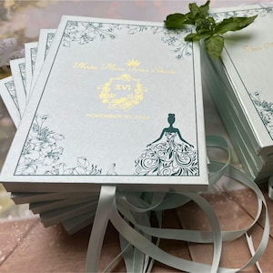 May include: A stack of light blue wedding invitations with gold foil text and floral accents. The invitations feature a silhouette of a woman in a gown and the date November 30, 2024. Light blue ribbons are tied around the stack.