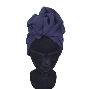Può includere: Un turbante blu navy con un grande accento in tessuto arricciato sulla parte superiore. Il copricapo è esposto su una testa di manichino nera su sfondo bianco. Il tessuto sembra morbido e fluido.