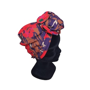 Könnte beinhalten: Ein farbenfrohes Kopftuch mit einem auffälligen Blumenmuster in Rot, Lila, Braun und Grün. Der Stoff ist oben gewickelt und geknotet, wodurch ein Turban-ähnlicher Stil entsteht. Dieses Accessoire verleiht jedem Outfit einen Farbtupfer.