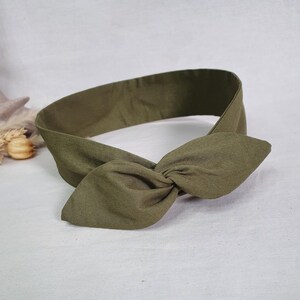 May include: Olive green headband with a knotted bow detail. The fabric appears to be a soft, textured material. The headband is designed to be worn around the head, with the bow positioned at the front.