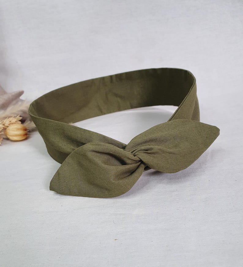 May include: Olive green headband with a knotted bow detail. The fabric appears to be a soft, textured material. The headband is designed to be worn around the head, with the bow positioned at the front.