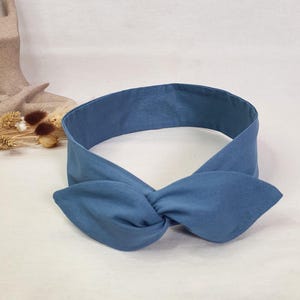 Könnte beinhalten: Ein blaues Stoff-Haarband mit geknoteter Schleife. Das Haarband ist einfarbig blau und hat eine dekorative Schleife auf der Vorderseite. Der Stoff wirkt weich und bequem.