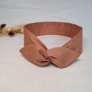 May include: A terracotta-colored headband with a knotted bow detail. The headband is made of a soft fabric and is designed to be worn around the head. The bow is centered on the front of the headband.