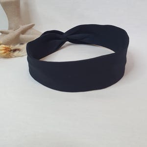 Puede incluir: Una diadema de tela negra con un dise&ntilde;o anudado. La diadema es de color liso y parece estar hecha de un material suave. La diadema se muestra sobre una superficie blanca.
