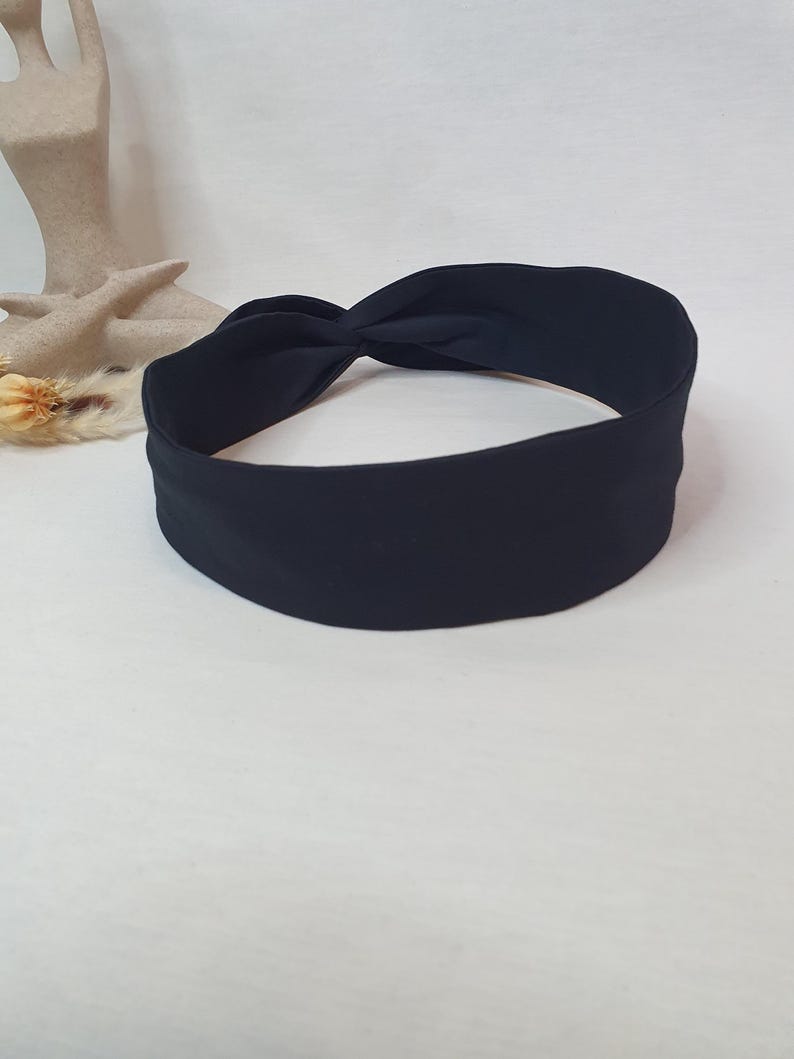 Puede incluir: Una diadema de tela negra con un dise&ntilde;o anudado. La diadema es de color liso y parece estar hecha de un material suave. La diadema se muestra sobre una superficie blanca.