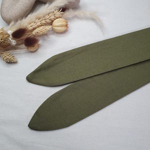 May include: Two olive green fabric headbands with pointed ends. The headbands are made of a soft, textured material. The background includes dried flowers and a neutral-colored sculpture.