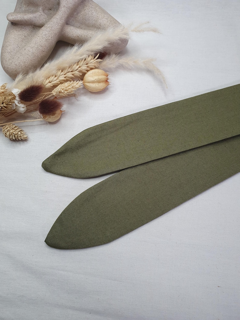 May include: Two olive green fabric headbands with pointed ends. The headbands are made of a soft, textured material. The background includes dried flowers and a neutral-colored sculpture.
