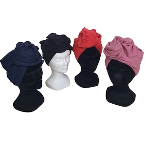Può includere: Quattro turbanti colorati esposti su teste di manichino. I turbanti sono blu navy, neri, rossi e malva. Ogni turbante presenta una grande rosetta decorativa sulla parte superiore. Le teste dei manichini sono nere e bianche.