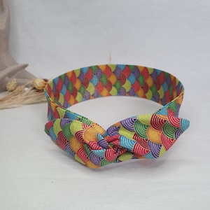 May include: A brightly coloured headband with a knotted bow. The fabric displays a repeating pattern of overlapping, semi-circular shapes in red, orange, yellow, green, blue, and purple. A vibrant hair accessory.