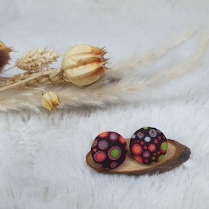 May include: Two button earrings with a dark background and a colorful polka dot pattern. The earrings are round and sit on a small, wooden slice. The earrings have red, green, and white dots.