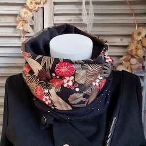 Puede incluir: Bufanda de cuello negra con un estampado floral y de pájaros en rojo, dorado y marrón. La bufanda se muestra sobre una cabeza de maniquí blanca y una chaqueta negra.