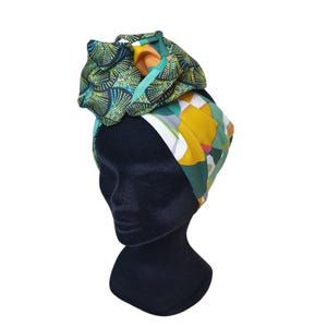 Op de afbeelding: Een kleurrijke tulband met een bloemenpatroon. De tulband heeft een mix van groene, gele en blauwe patronen. De tulband wordt gepresenteerd op een zwarte mannequinhoofd.