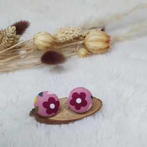 May include: Two round, pink fabric earrings with a floral pattern in white and burgundy. The earrings are displayed on a small, natural wood slice, with dried flowers and fluffy white fabric in the background.