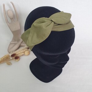 May include: An olive green headband with a knotted bow detail. The headband is displayed on a black mannequin head. The fabric appears to be a soft, textured material. The background is a neutral white.