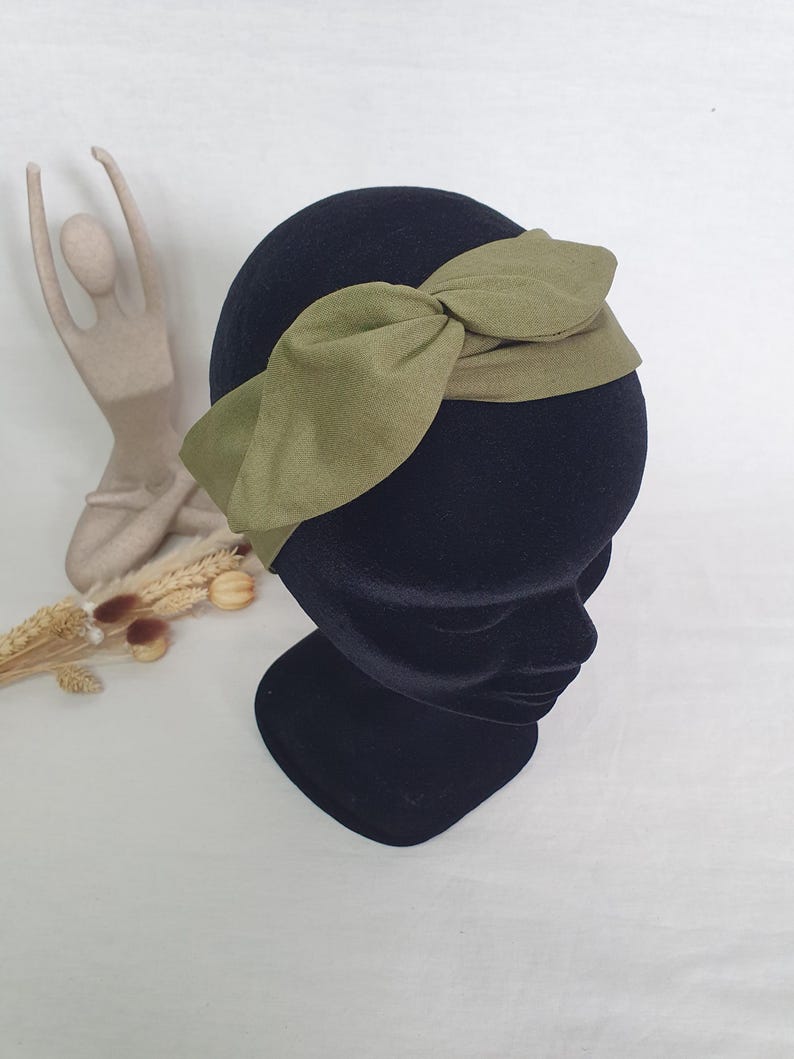 May include: An olive green headband with a knotted bow detail. The headband is displayed on a black mannequin head. The fabric appears to be a soft, textured material. The background is a neutral white.