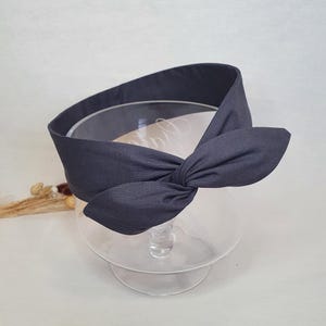 May include: A dark gray fabric headband with a knotted bow detail. The headband is made of a soft material and is designed to be worn around the head. The bow is centered on the top of the headband.