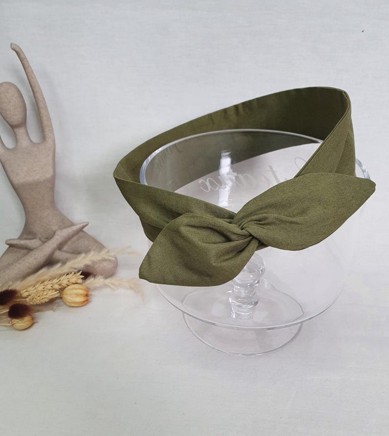 May include: An olive green headband with a knotted bow detail. The headband is made of fabric and is displayed on a clear glass pedestal. A decorative statue and dried flowers are in the background.