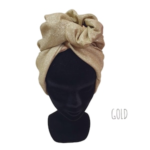 Può includere: Un turbante dorato con un tessuto strutturato e scintillante è esposto su una testa di manichino nera. Il turbante è annodato nella parte superiore, creando un design elegante e voluminoso. La parola "GOLD" è scritta in un carattere scritto a mano.