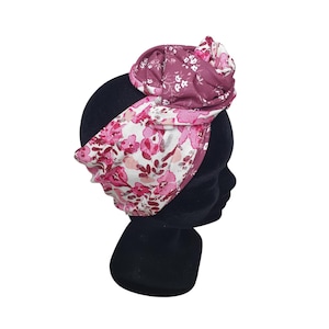 May include: A floral headband with a knotted top detail. The headband features a pattern of pink flowers on a white background, with a solid burgundy fabric on the top. The headband is displayed on a black mannequin head.